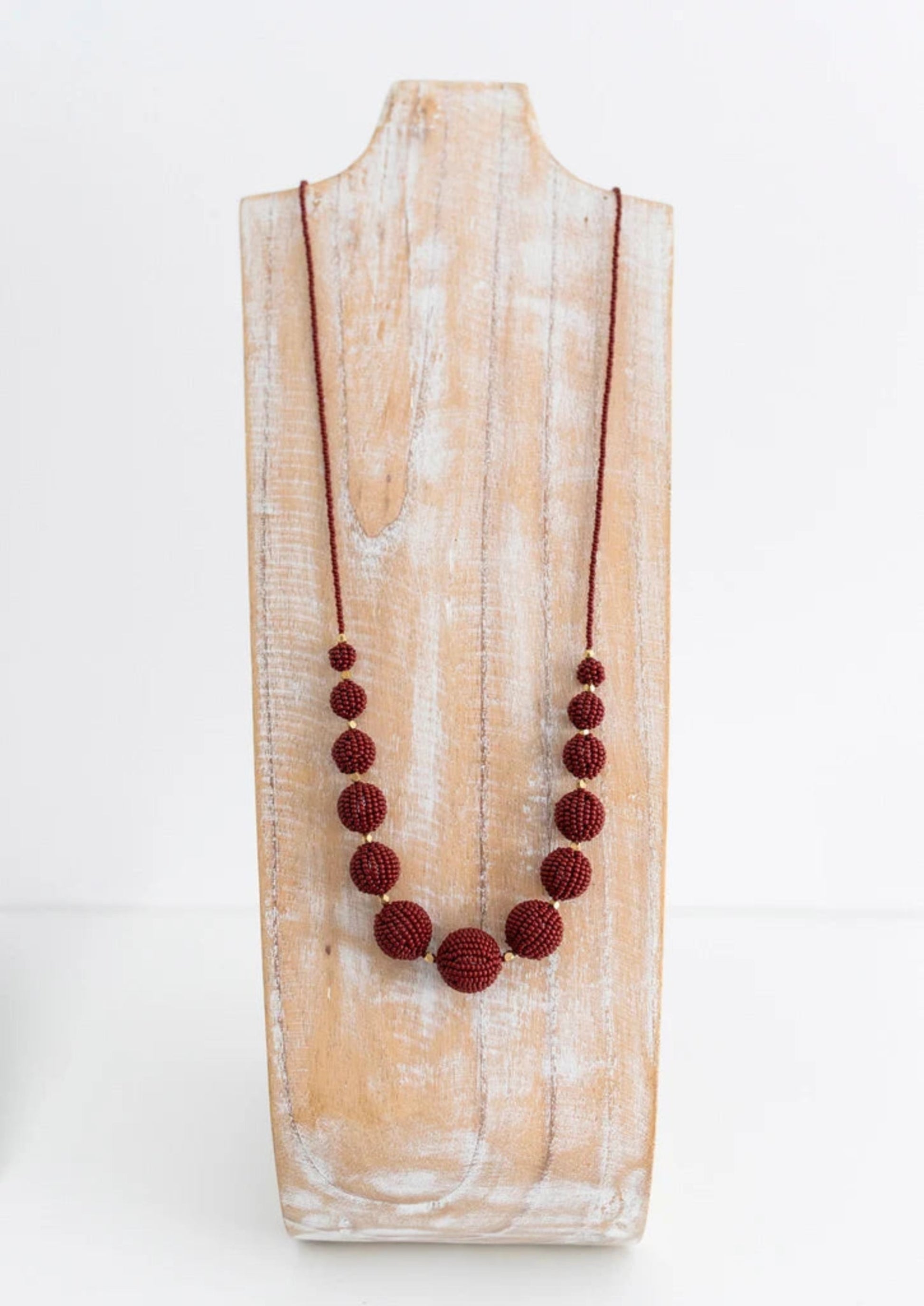 A dark red beaded necklace with textured round beads and a cord chain, displayed on a rustic white-washed wooden stand against a white background.