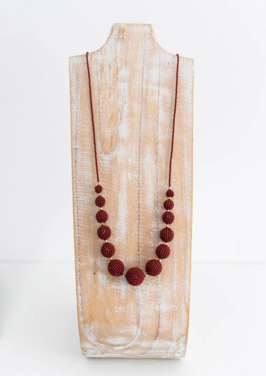 A dark red beaded necklace with textured round beads and a cord chain, displayed on a rustic white-washed wooden stand against a white background.